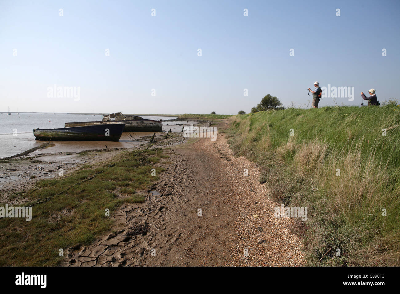 River Ore / Alde, bank / wall at Orford Suffolkl, UK abandoned boats in background with tourists ramblers taking photographs Stock Photo