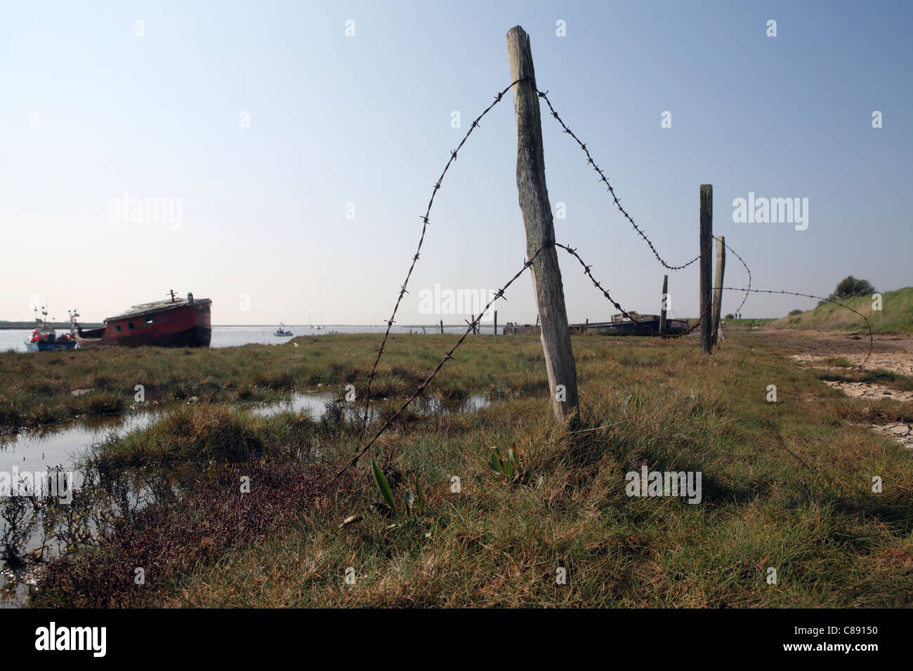 River Ore / Alde, bank / wall at Orford Suffolk, UK showing abandoned boats in background and barbed wire in foreground Stock Photo