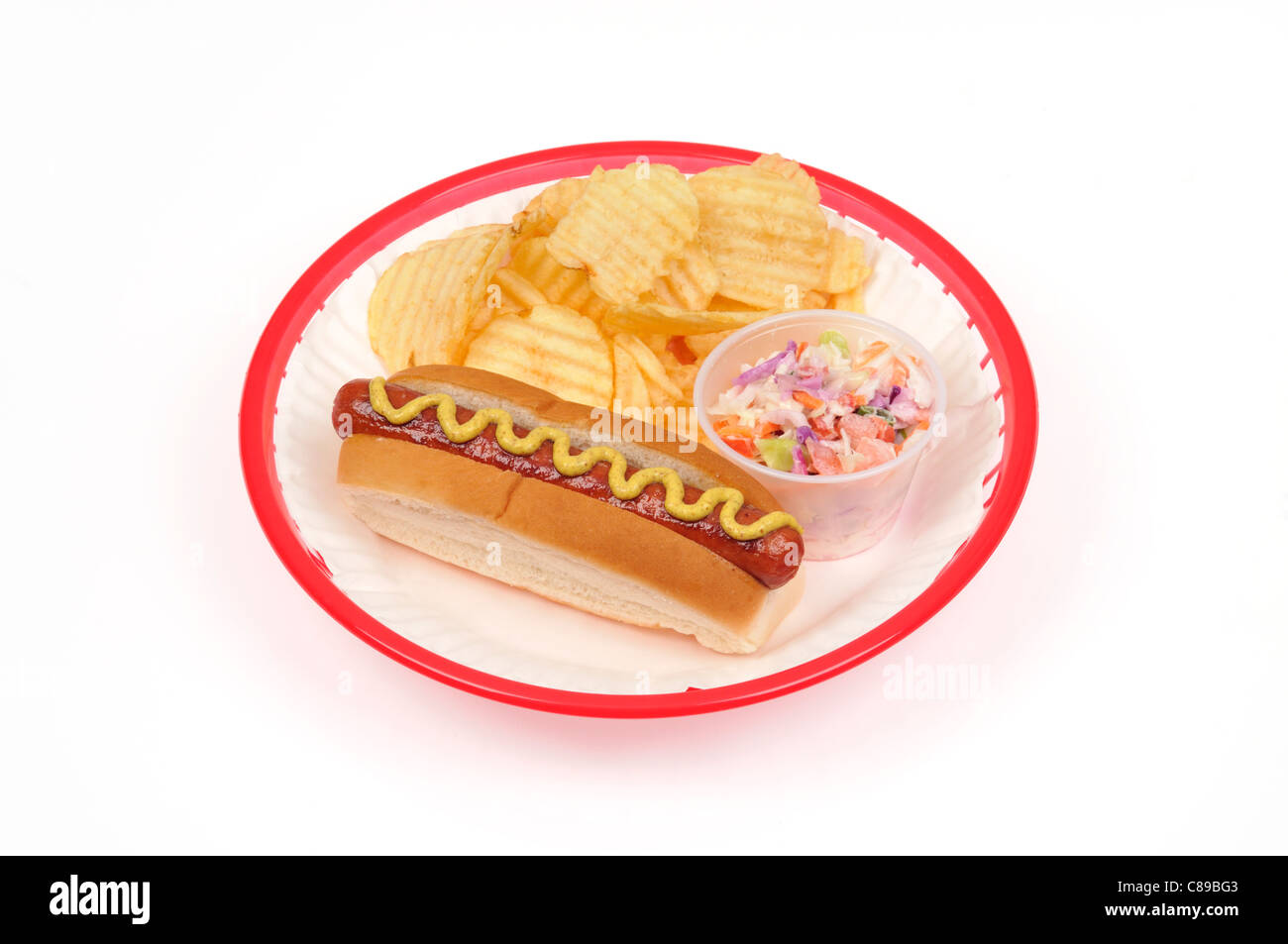 Hotdog with mustard, crisps or potato chips and coleslaw on white paper plate in red plastic basket on white background cutout. Stock Photo