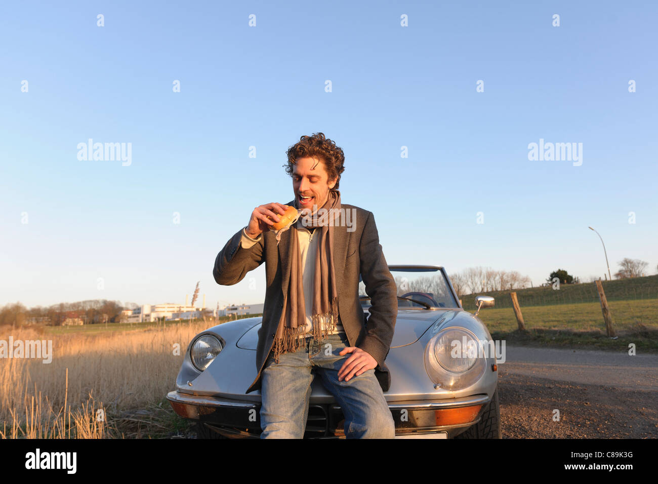 Germany, Hamburg, Man eating fish sandwich beside classic cabriolet car Stock Photo