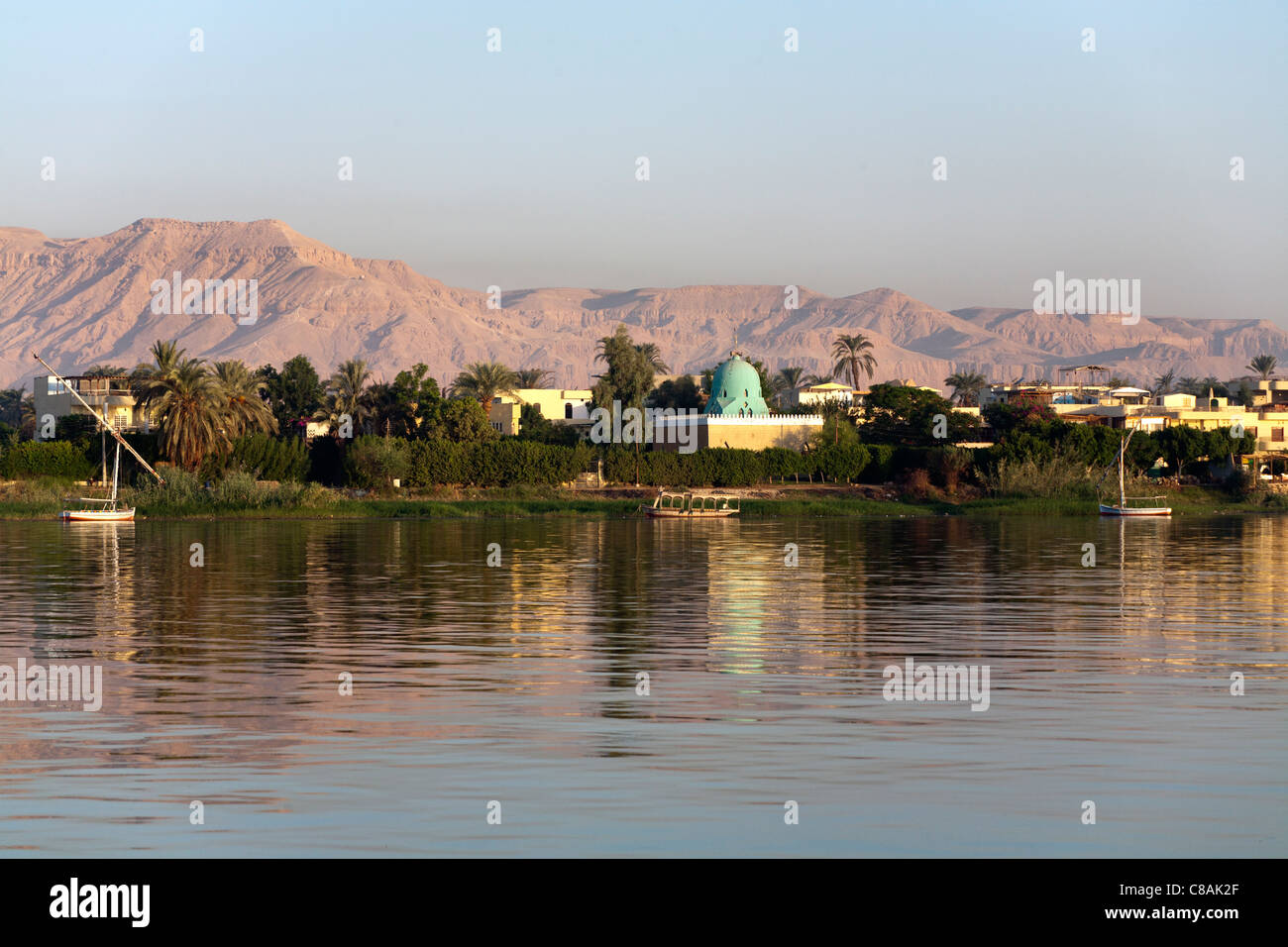 Section of Nile river bank with trees and mosque and the West Bank mountains in the background lit by golden evening light Stock Photo