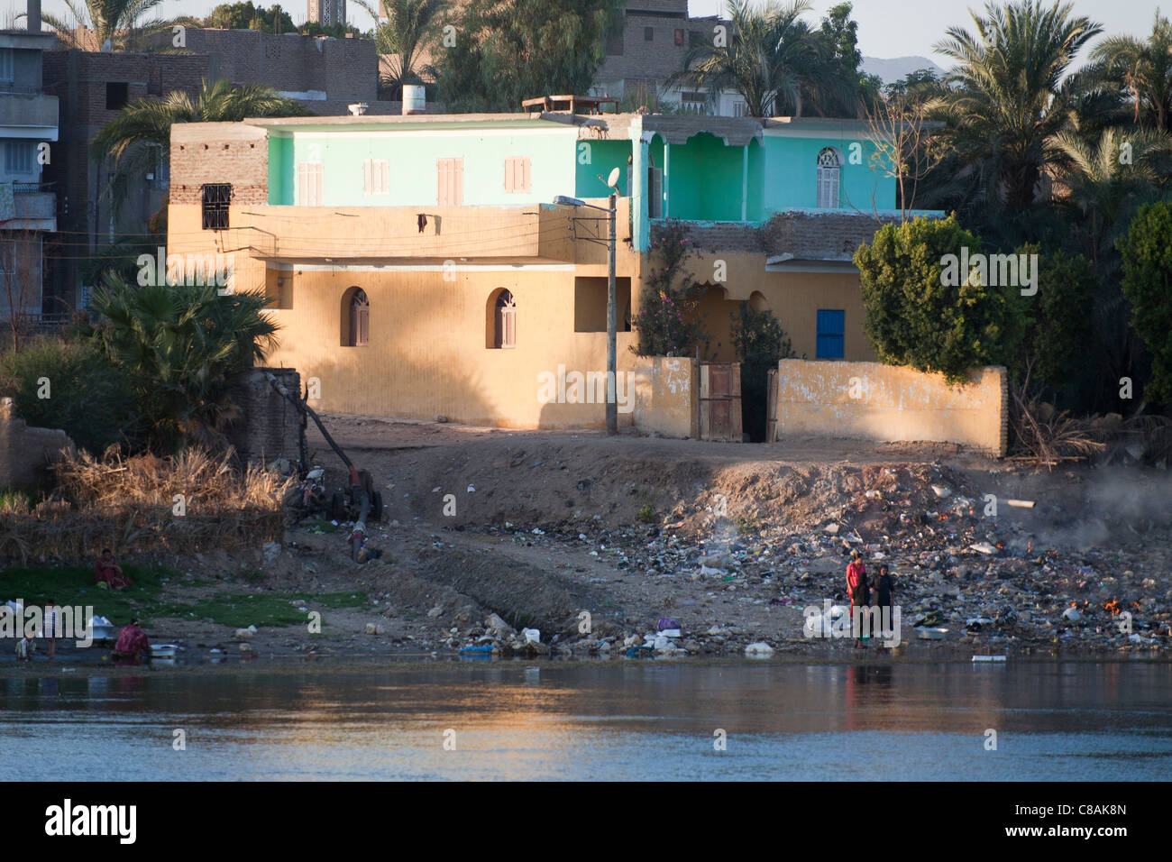 Traditional Egyptian house on the Nile bank with palm trees and rubbish covered bank and two women talking, Egypt Stock Photo