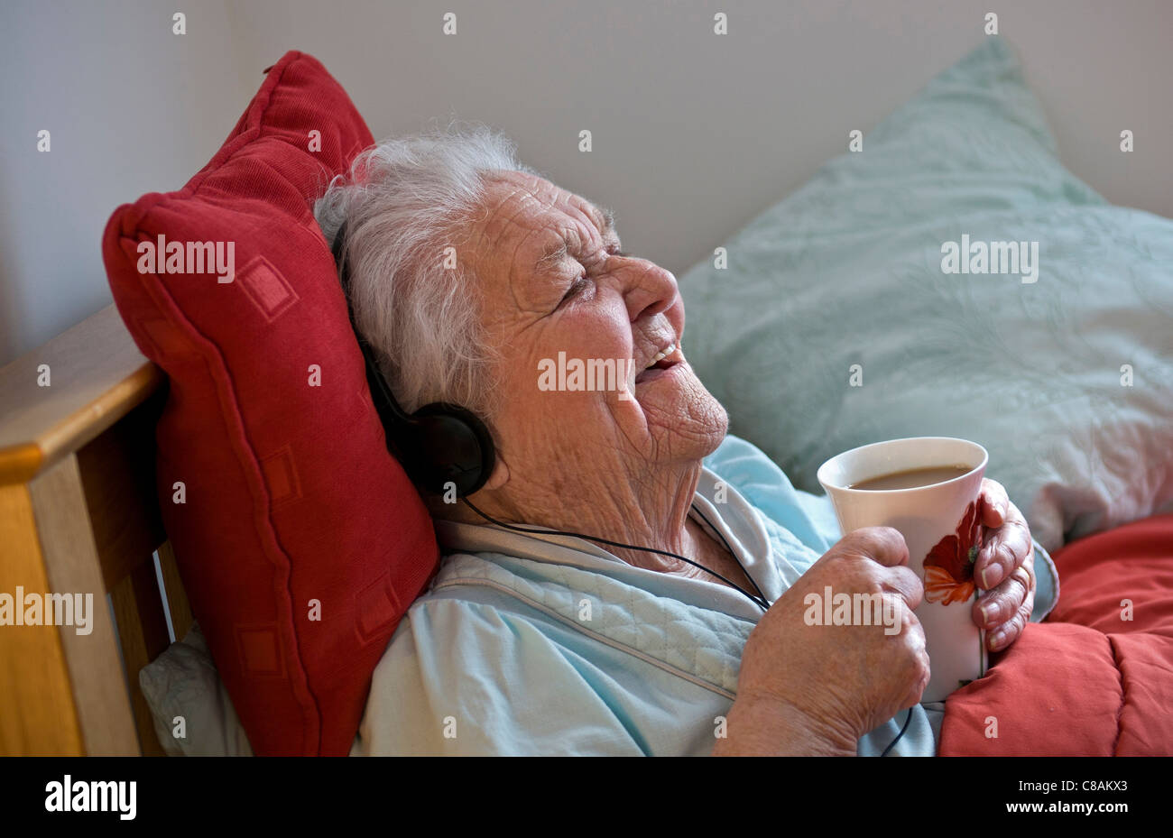 Happy contented elderly  lady  resting in bed wearing headphones enjoys a cup of tea in her sunny comfortable room Stock Photo