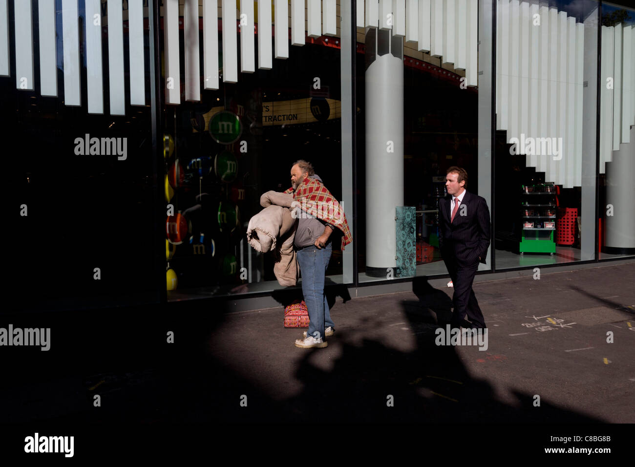 A smart suited businessman walks past an unfortunate homeless man holding his worldly possessions. Stock Photo