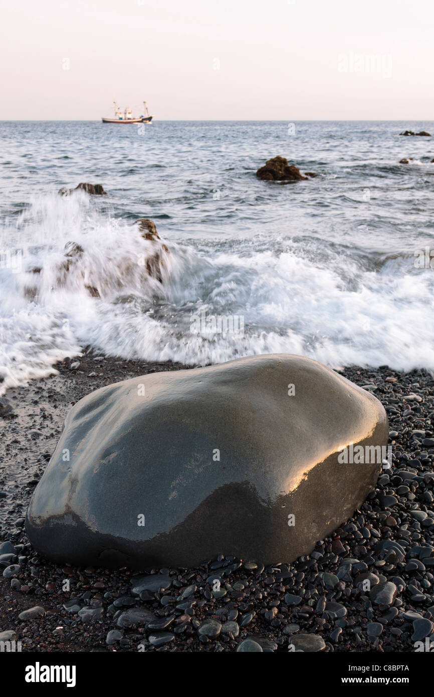 Smooth rock on the shore as a fishing boat sets off at sunset on the ...