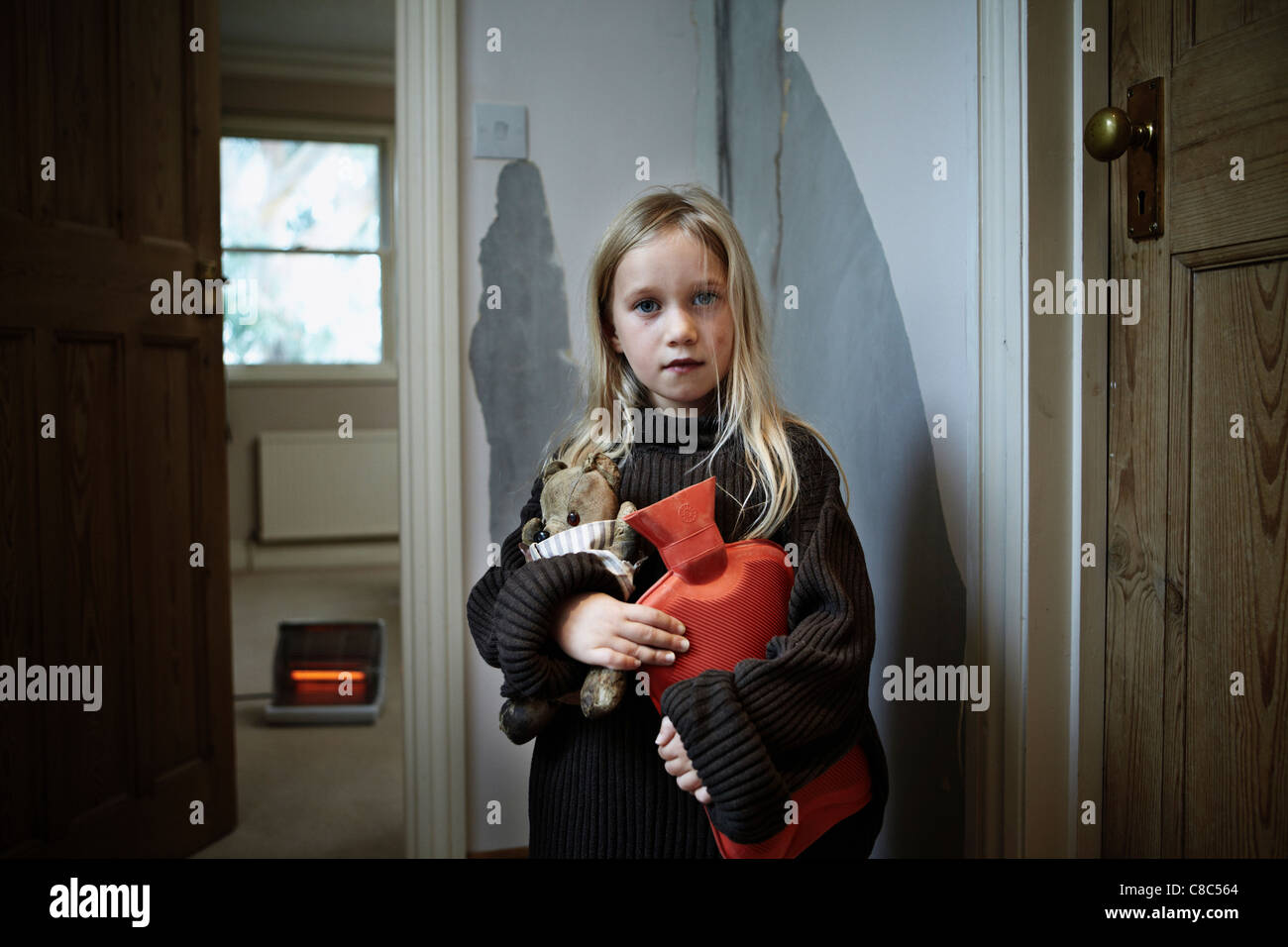 A  young girl holding a hot water bottle and wearing a large jumper in a run down flat with a electric fire in back ground Stock Photo