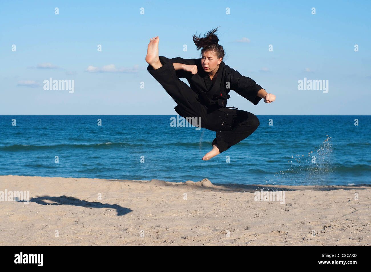 A female, fourth degree, Taekwondo black belt athlete performs a midair jumping kick on the beach. Stock Photo