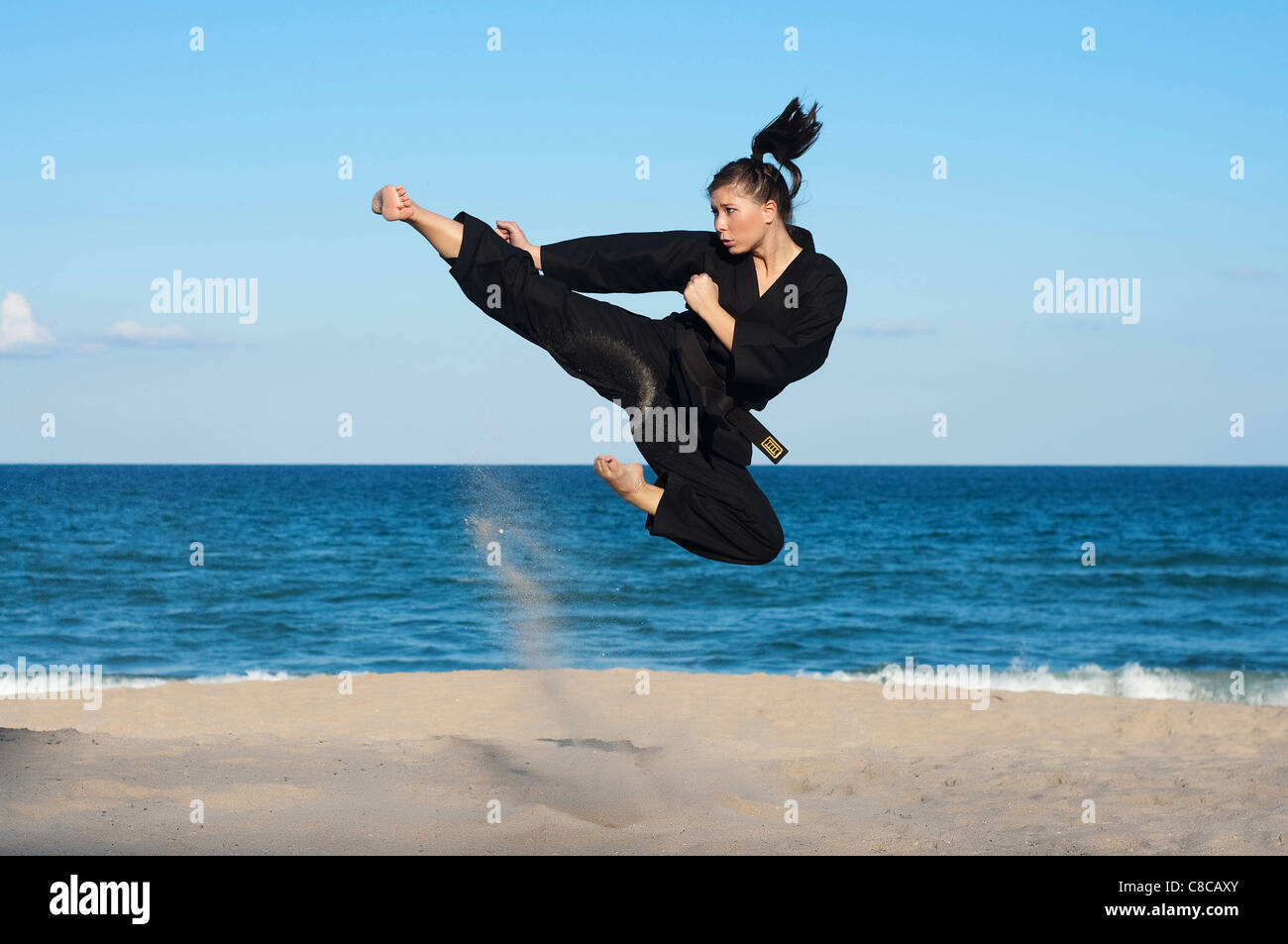 A female, fourth degree, Taekwondo black belt athlete performs a midair jumping kick on the beach. Stock Photo