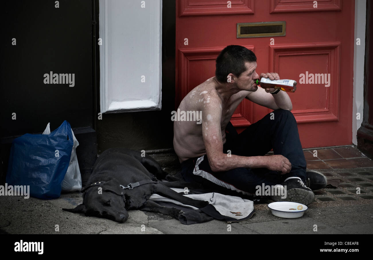 Homeless man with his dog sitting with begging bowl on shopping center high street drinking spirit alcohol ignored by passers by Stock Photo