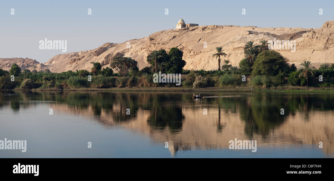 Section of Nile river bank with five men fishing in small boat with trees, a hill with a shrine on top, and perfect reflection Stock Photo