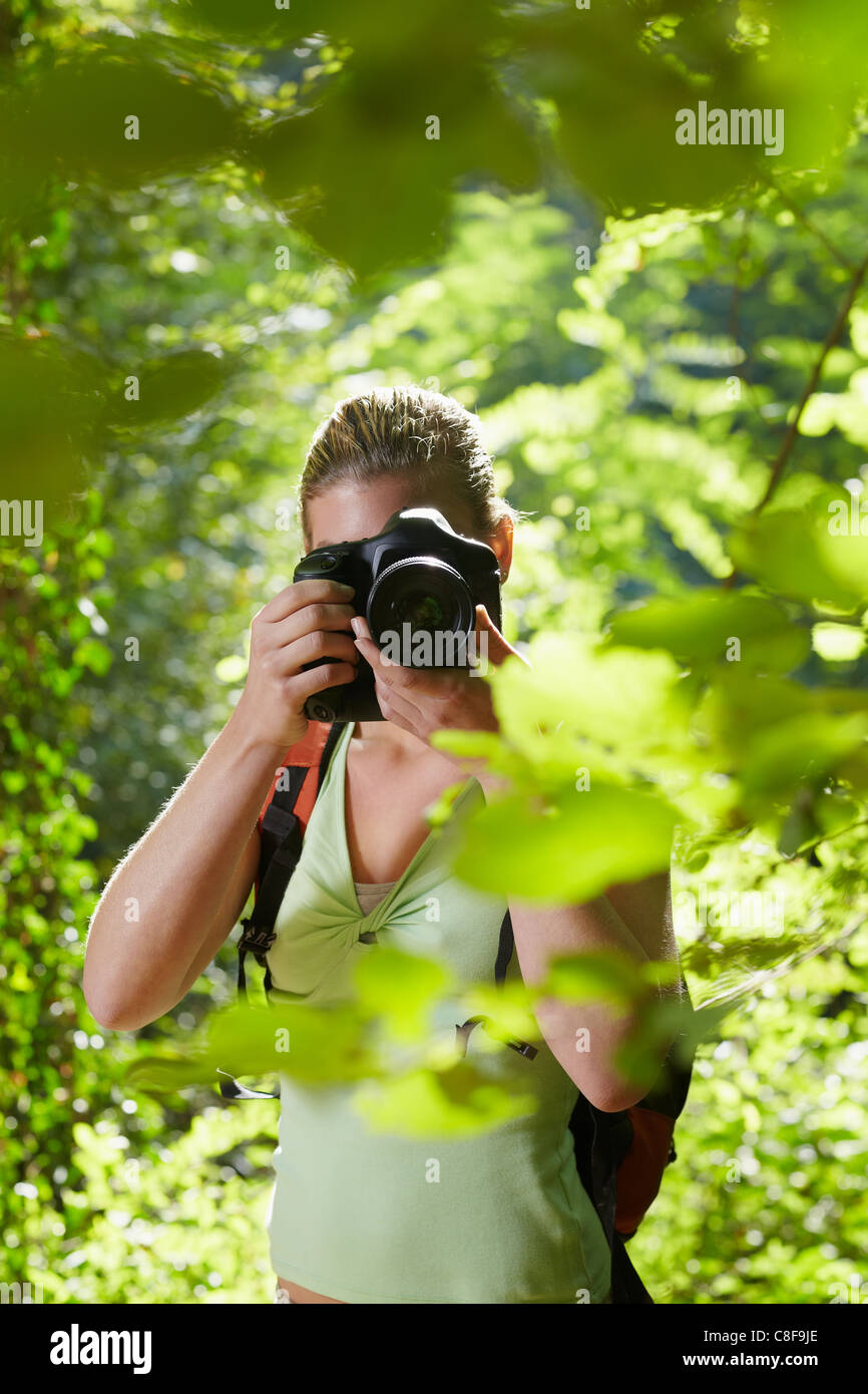 young woman trekking among trees and taking pictures with dslr camera. Vertical shape, front view, waist up Stock Photo