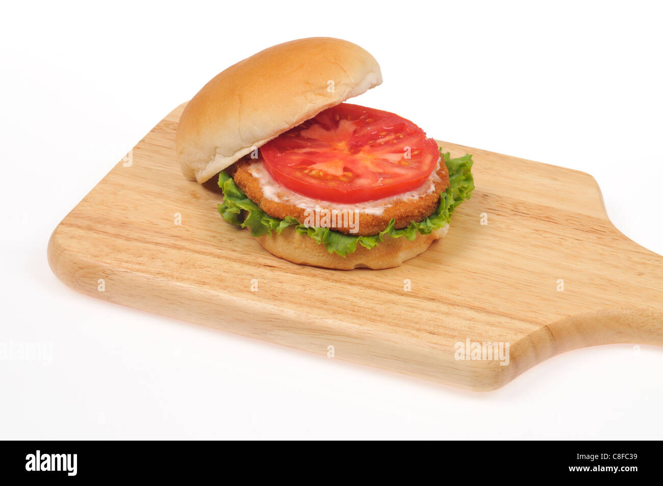 Chicken burger breaded patty with lettuce and tomato and mayonnaise on bread roll on wood cutting board on white background. Stock Photo