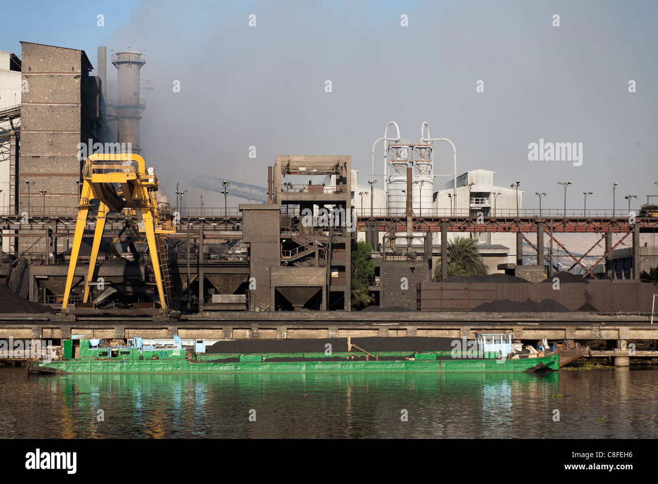 A section of Nile river bank with industrial plant belching smoke and coal barge moored at the quay in front reflected in water Stock Photo