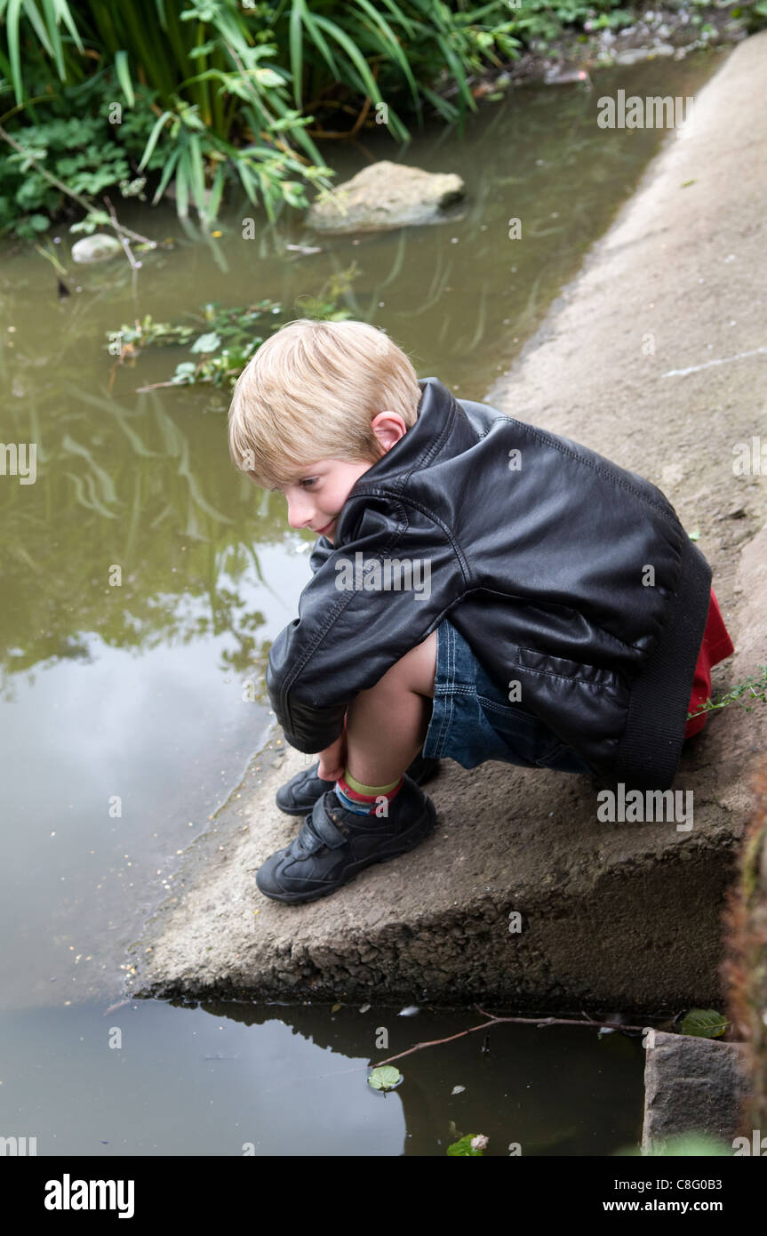 8 year old Caucasian boy sat watching at a pond taken at Barrs Court Moat Pond, near Longwell Green in Bristol, UK Stock Photo