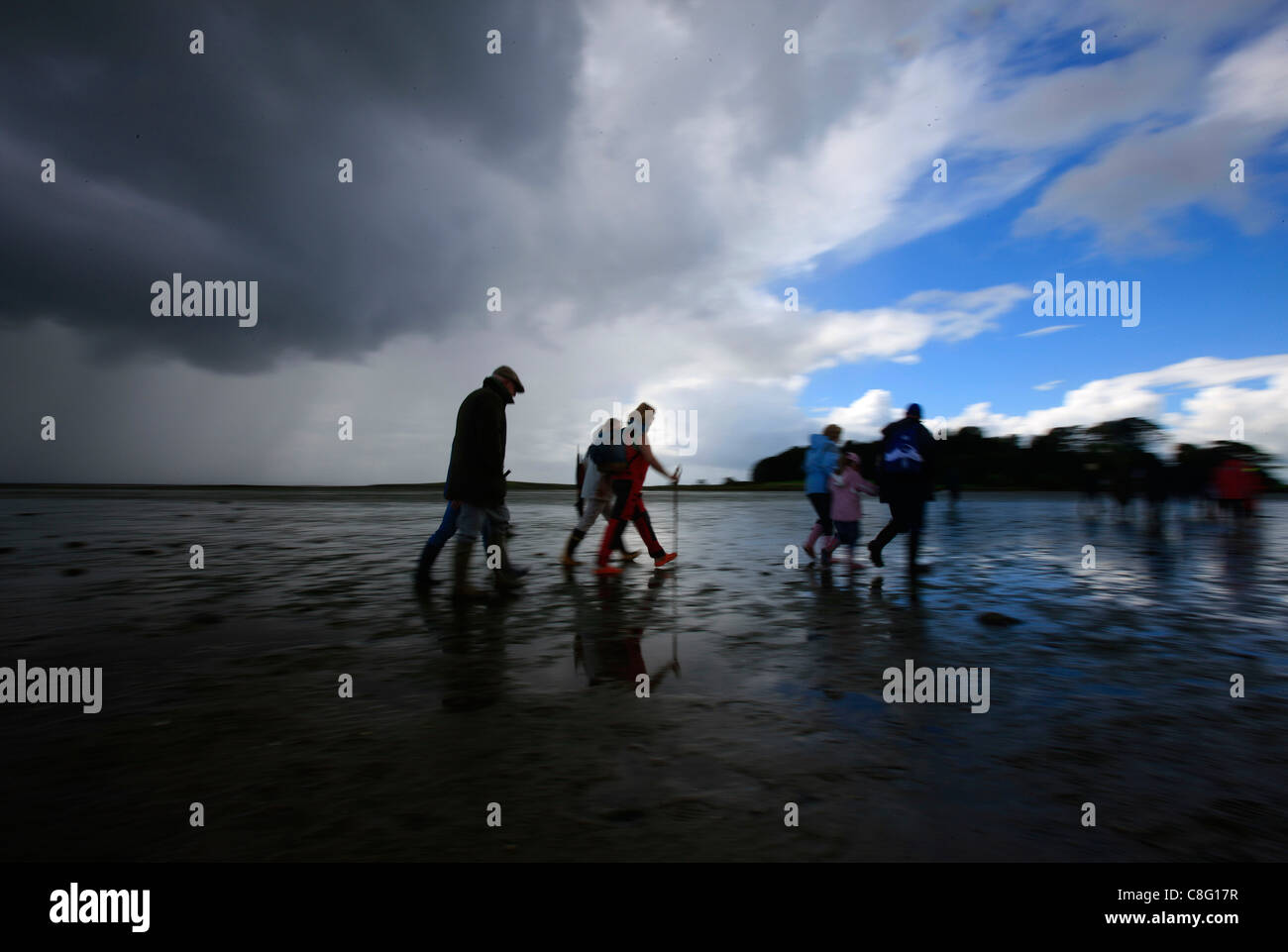 A small group of people explore as they walk along the shore under a stormy sky Stock Photo