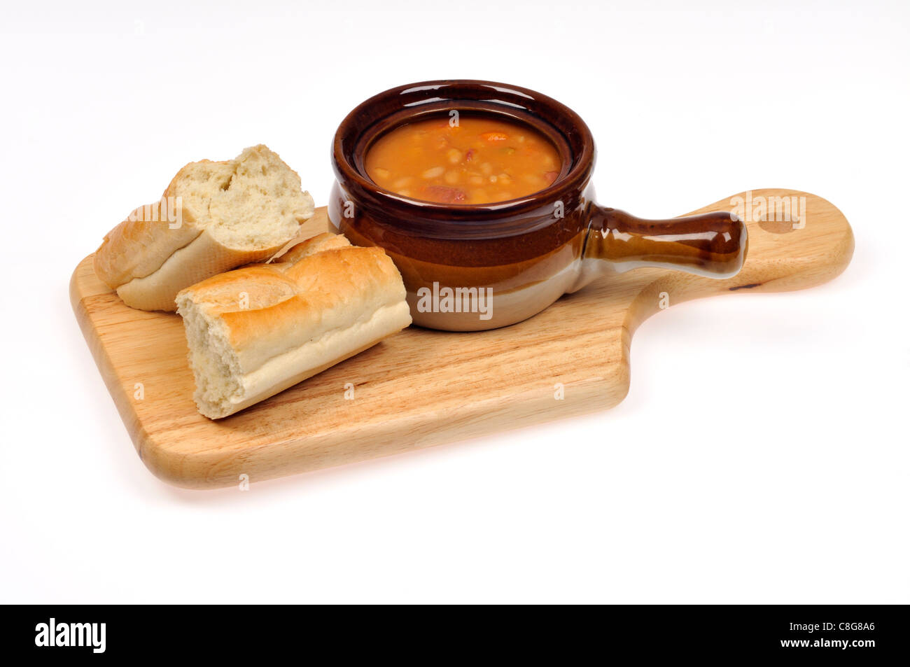 Ham & bean soup in a soup crock with french bread on wood cutting board on white background. Stock Photo