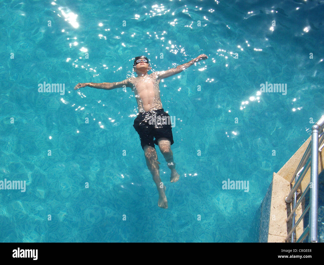 A young boy floating on his back in a swimming pool with sun reflecting ...