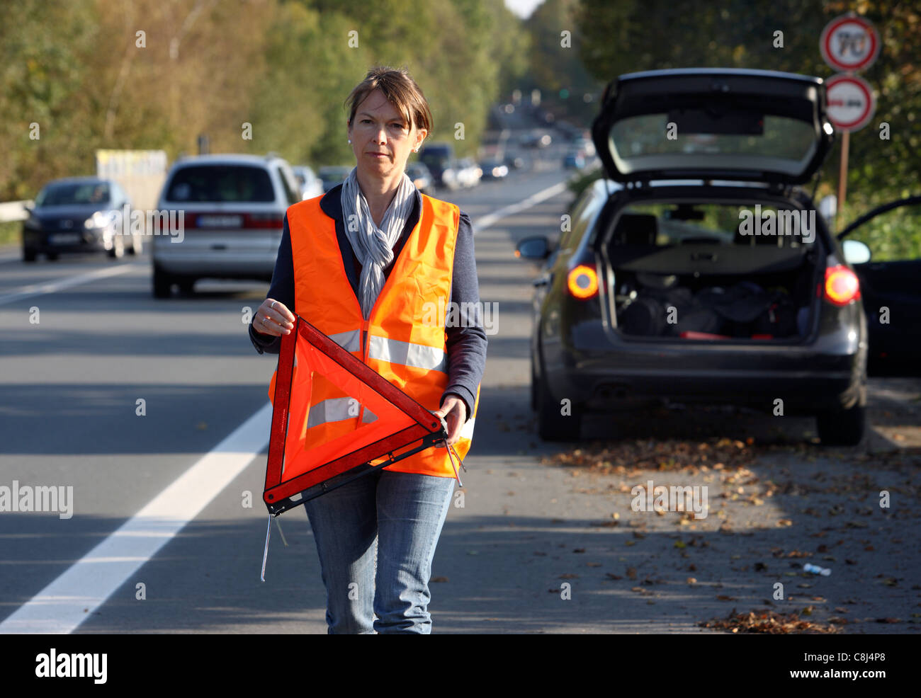 Car breakdown, on a highway, female driver, wearing a high visibility vest,  set up a warning triangle. Stock Photo