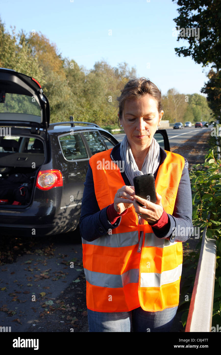 Car breakdown on a highway, woman, female driver, wearing a high visibility vest, calling for help with her mobile phone. Stock Photo