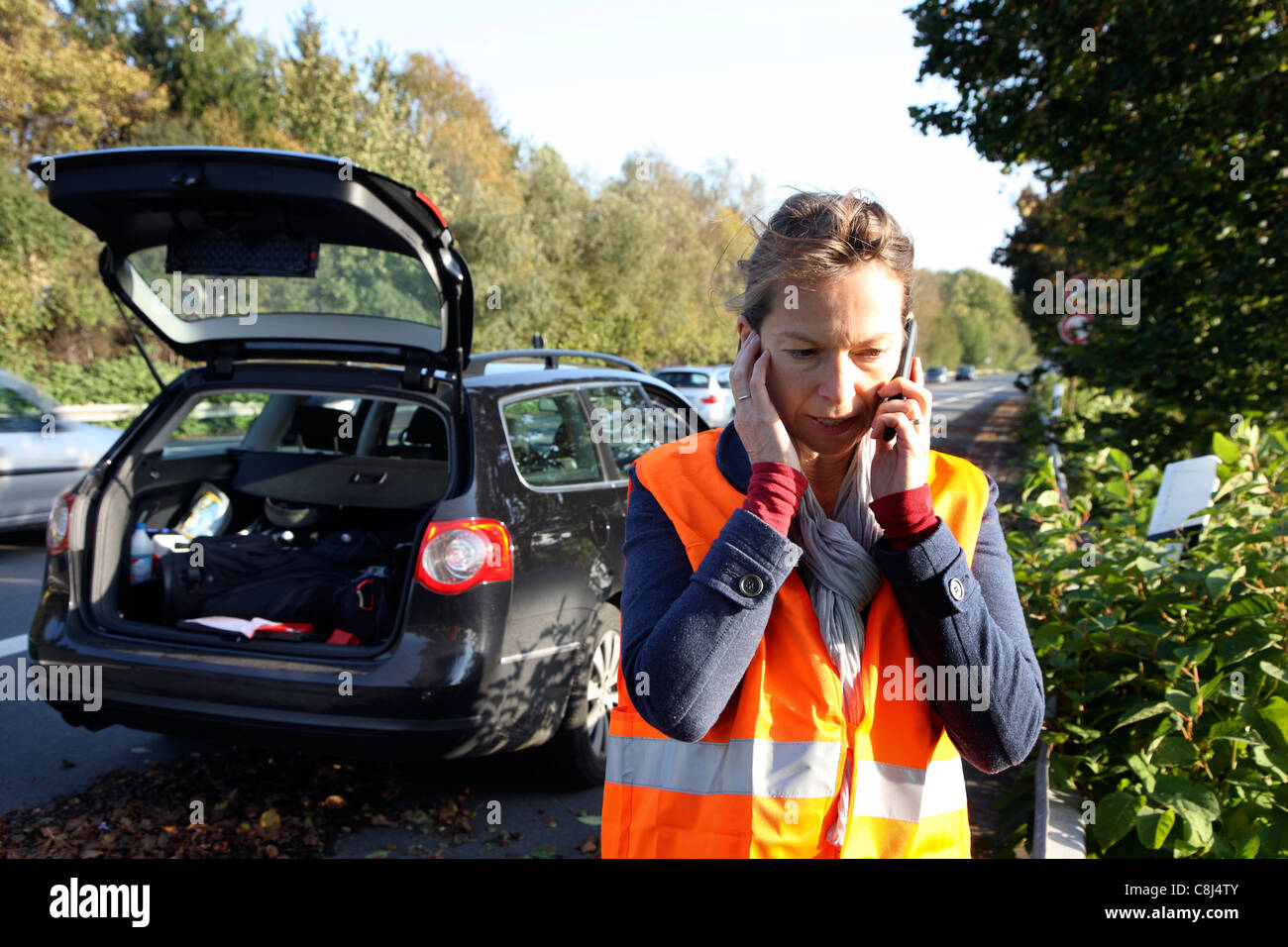 Car breakdown on a highway, woman, female driver, wearing a high visibility vest, calling for help with her mobile phone. Stock Photo