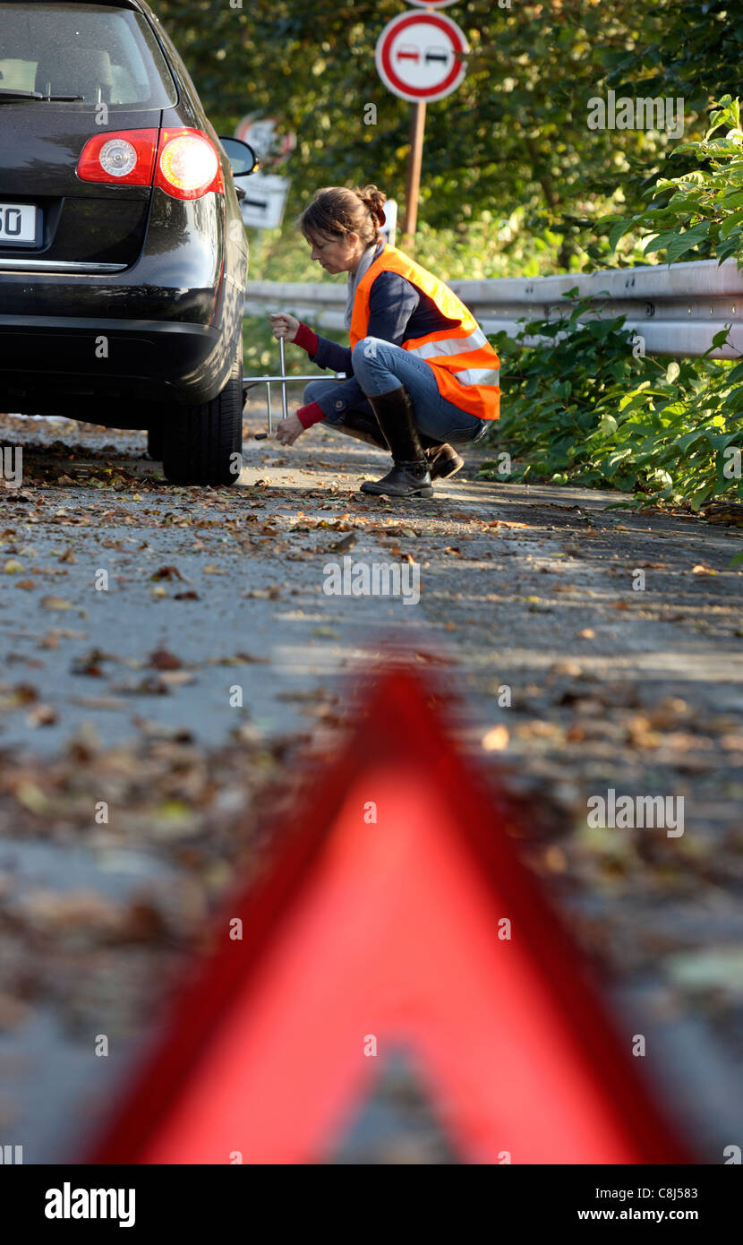 Car breakdown, flat tire. Woman changes a tire of a car on a highway, wearing a high visibility vest. Stock Photo