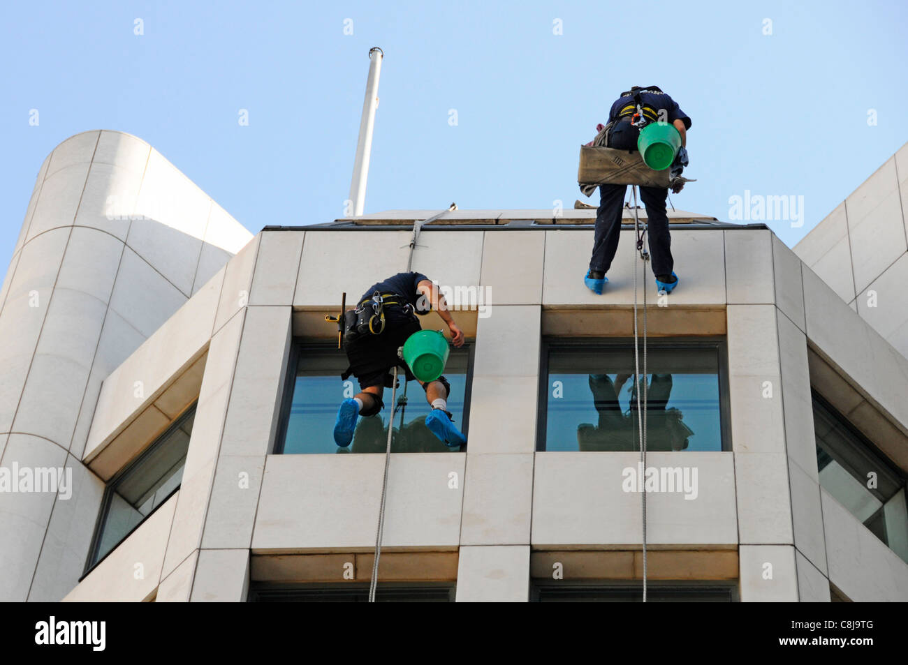 Window cleaner workers abseiling from roof level down face of office block reflections in glass London England UK Stock Photo