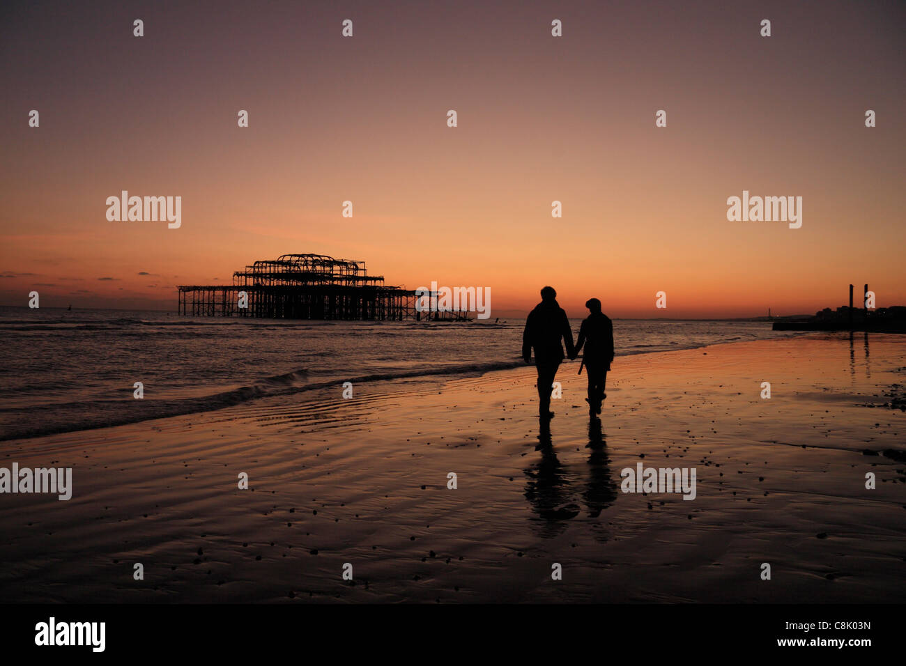 A couple walking hand in hand along Brighton beach, East Sussex, UK  towards the ruins of the West Pier. Stock Photo