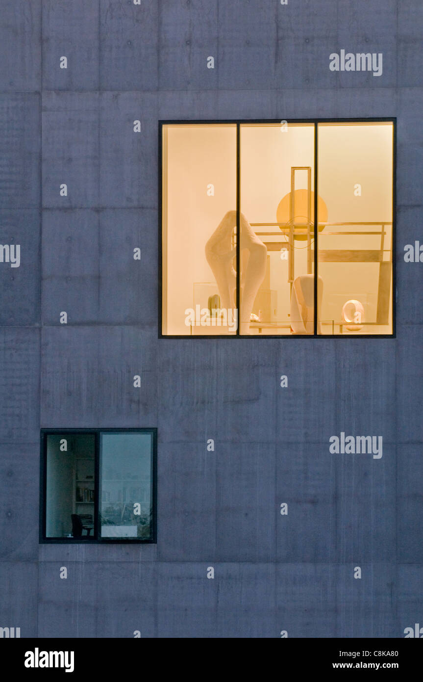 View through large window of Hepworth Gallery, lights on (golden glow) & sculptural pieces exhibited inside - Wakefield, West Yorkshire, England, UK. Stock Photo