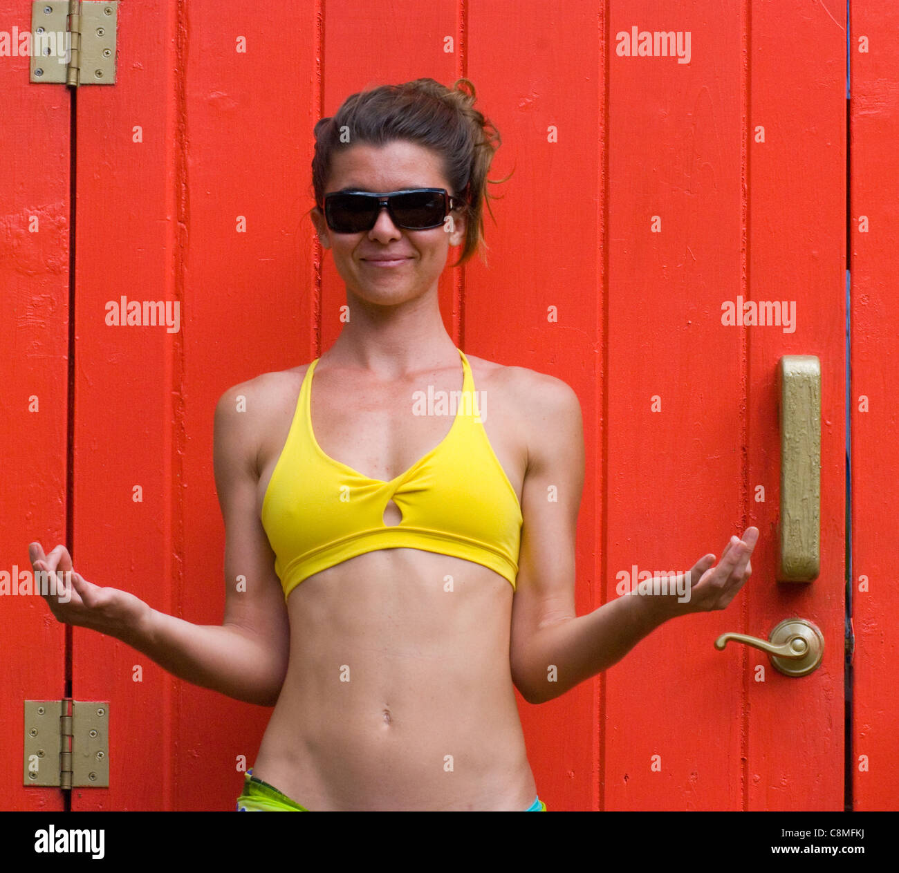 Young woman, age 21, in sunglasses standing in yellow swim suit in front of bright orange door making a mudra with her hands Stock Photo