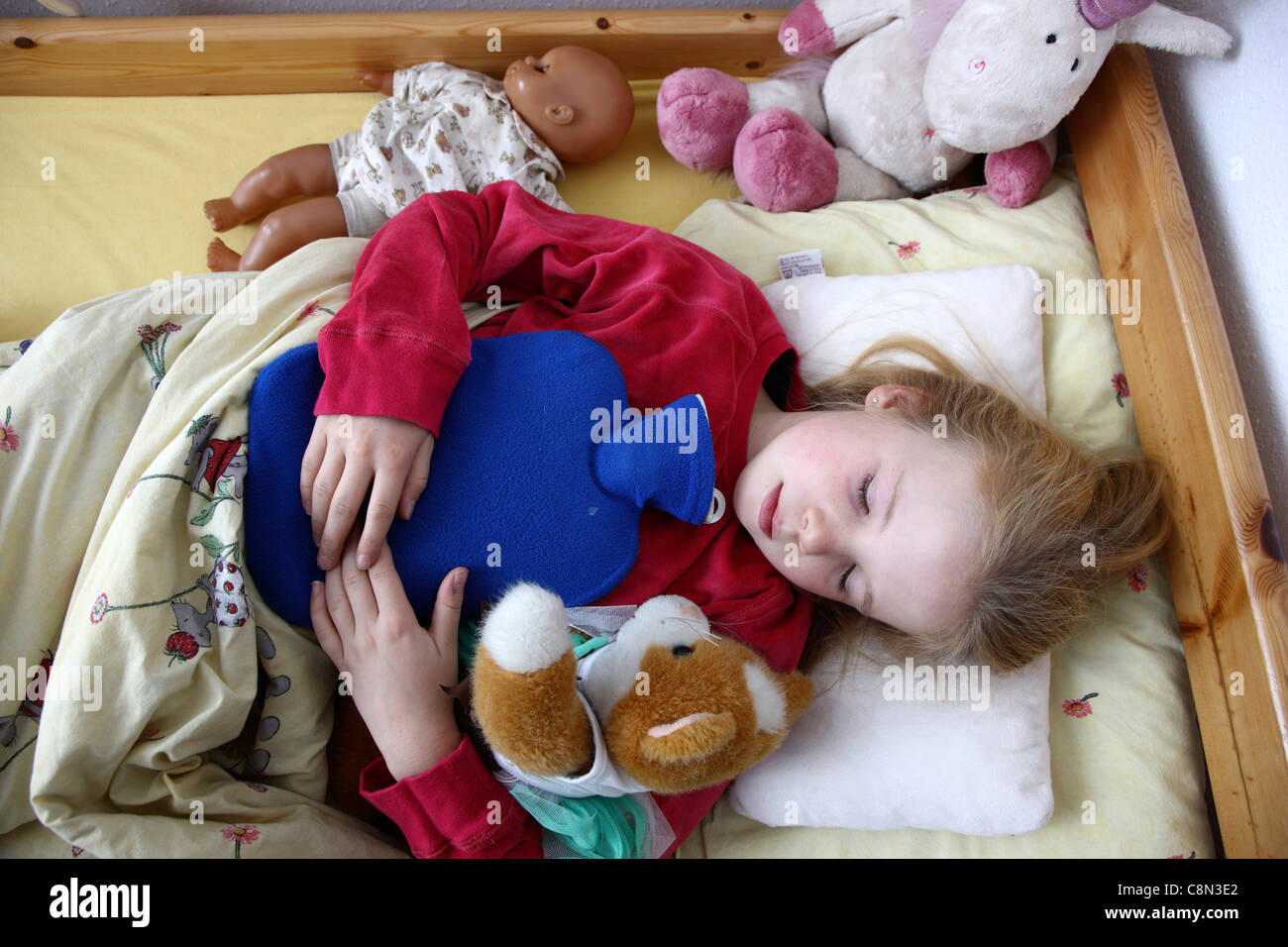 Young girl, 10 years old, lies in bed, at home, sick with a cold. Using a hot-water bag, accompanied by some cuddly pets. Stock Photo