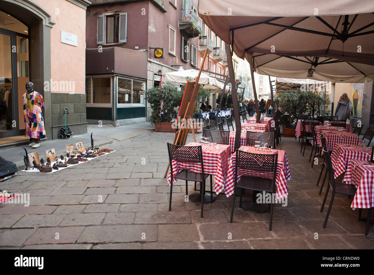 Outdoor restaurant in the Brera district of Milan. African vendor selling items in front of the tables. Italy Stock Photo