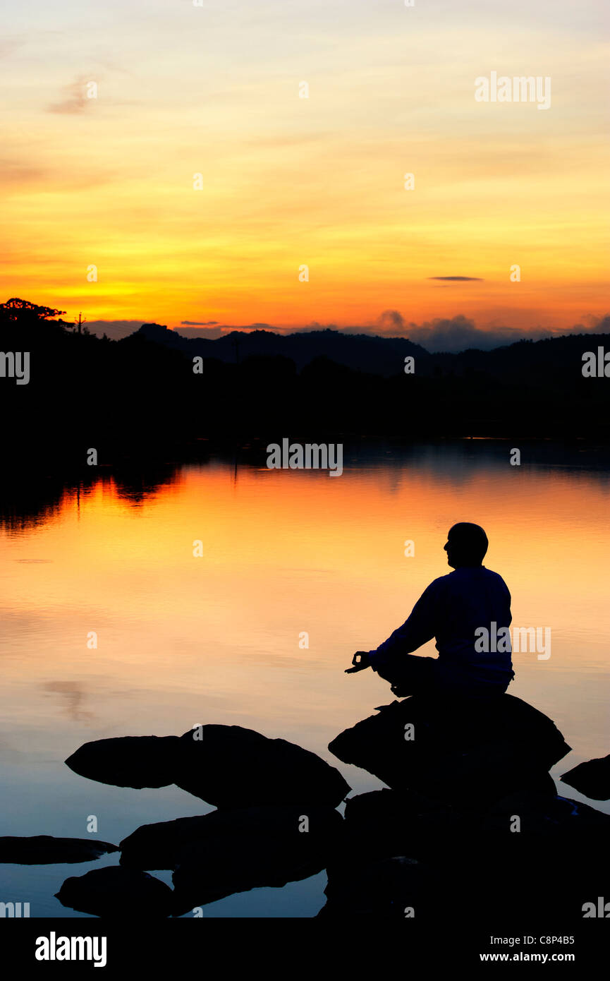 Sunrise silhouette of a man meditating on rocks at an Indian lake ...