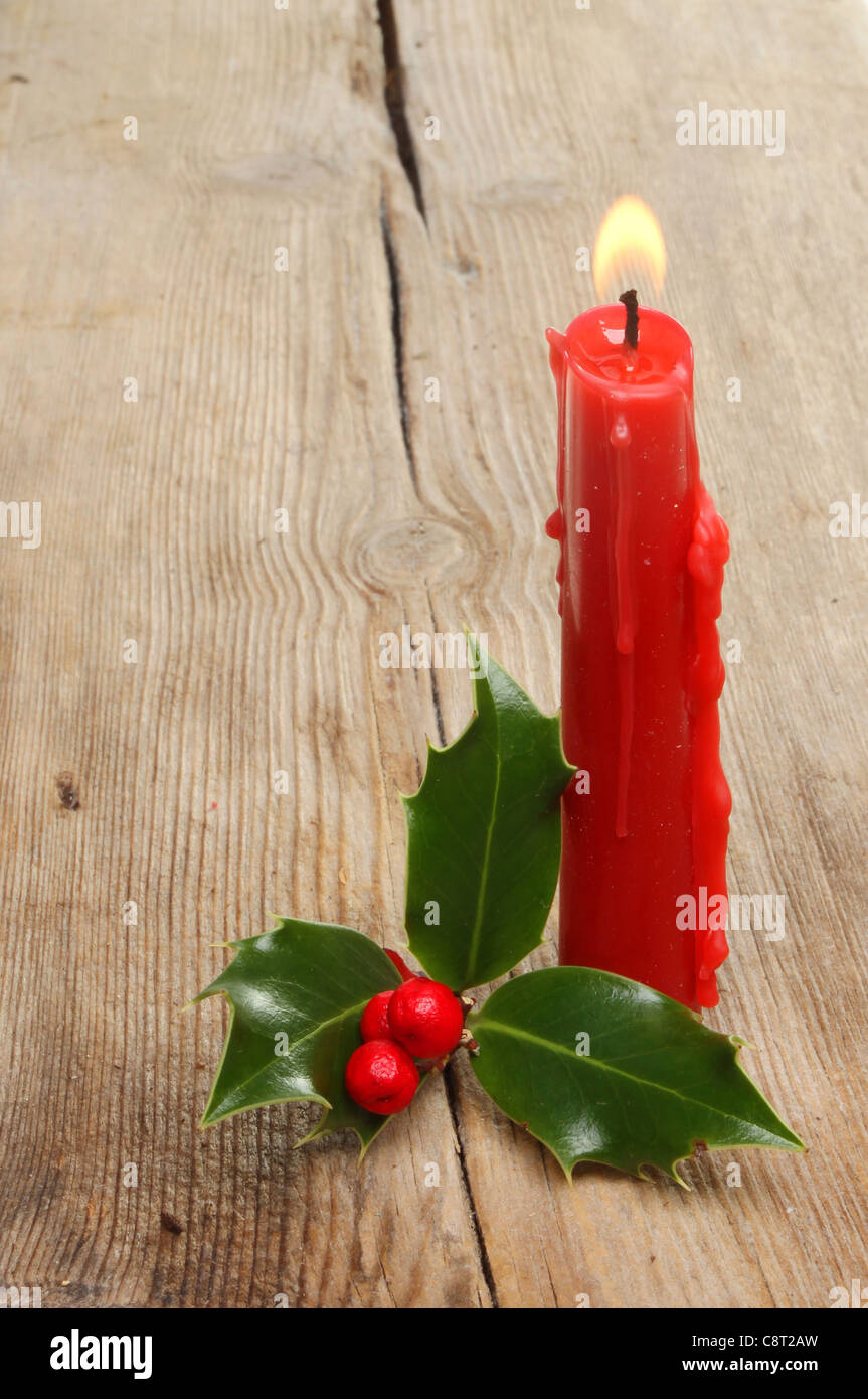 Burning red candle and a sprig of holly with red berries on old weathered wood Stock Photo