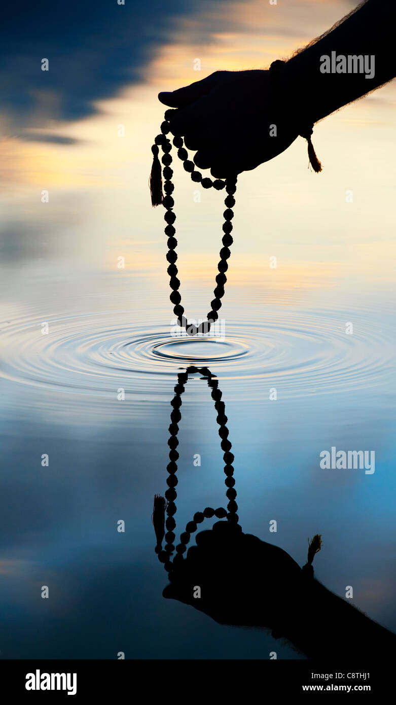 Hand holding Indian Rudraksha / Japa Mala prayer beads over rippling water . Silhouette Stock Photo