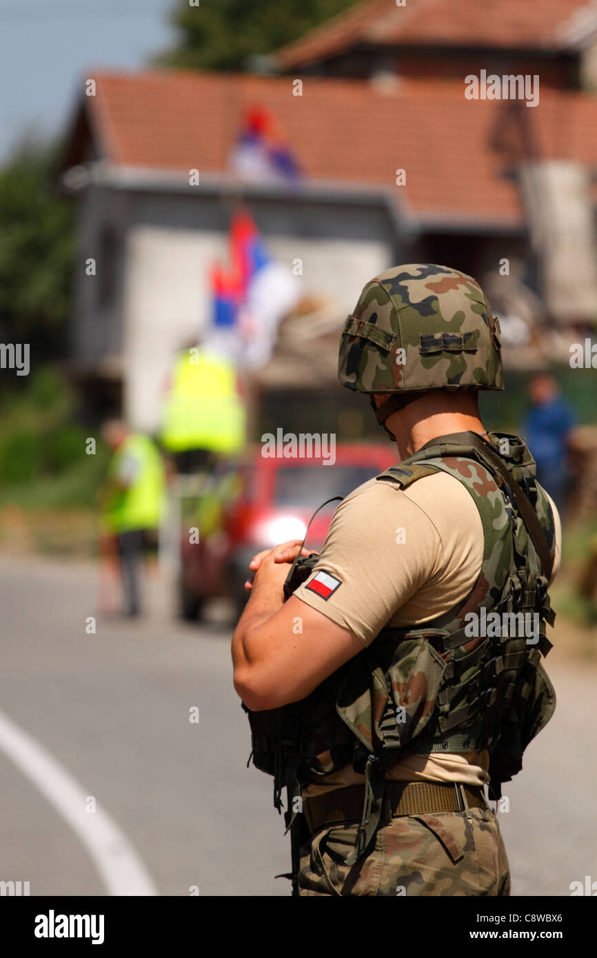 Polish KFOR soldier in combat gear on a road block in Kosovo, Serb flags in background Stock Photo