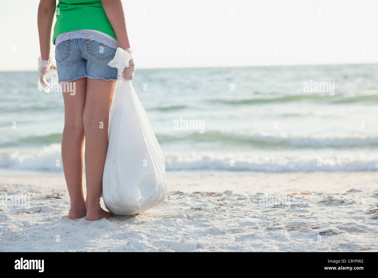 USA, Florida, St. Petersburg, Rear view of girl (10-11) standing on beach with garbage bag, facing ocean Stock Photo