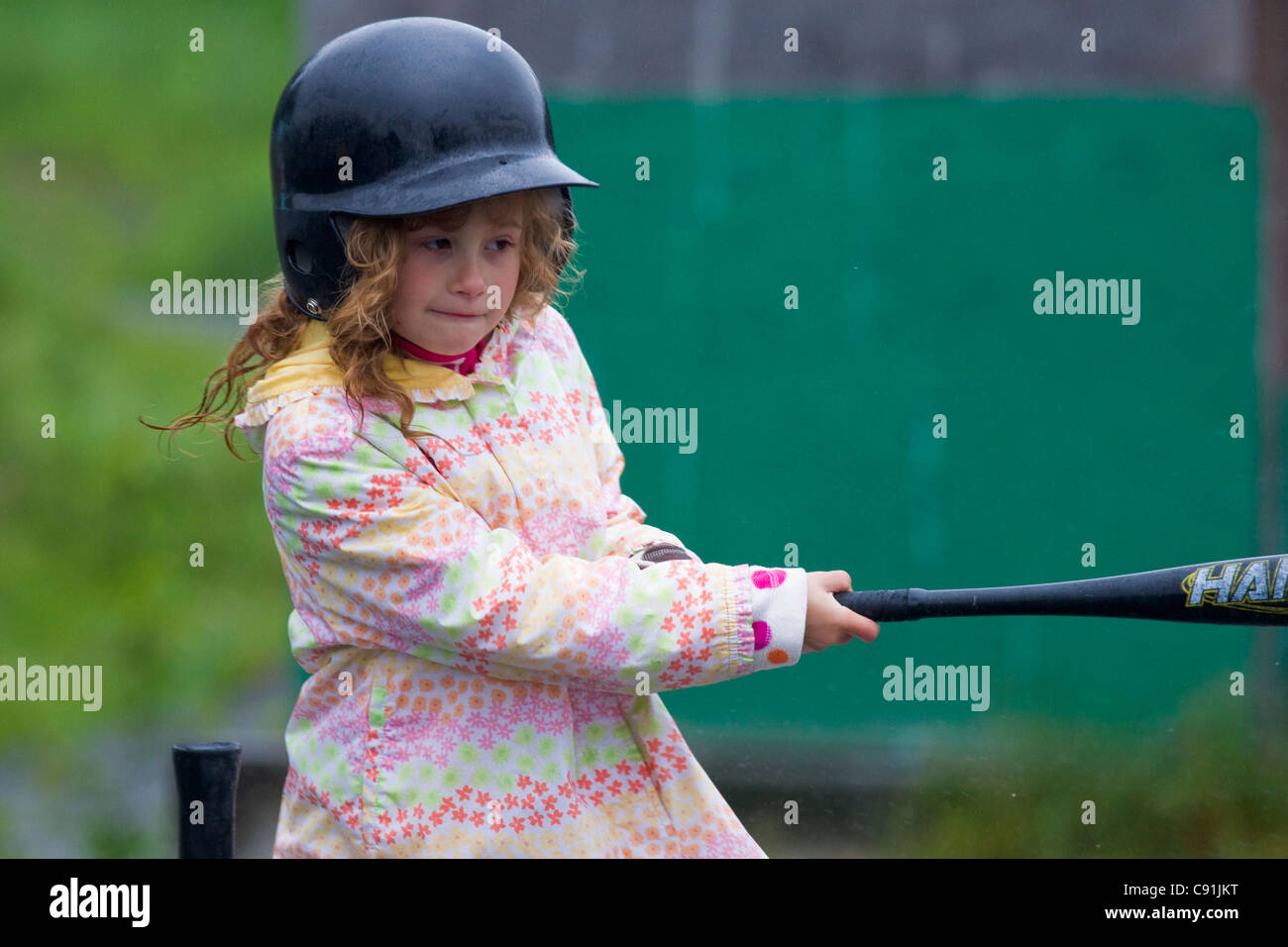 Young girl swinging at T-ball while wearing rain coat, Cordova little league, Cordova, Southcentral Alaska, Summer Stock Photo