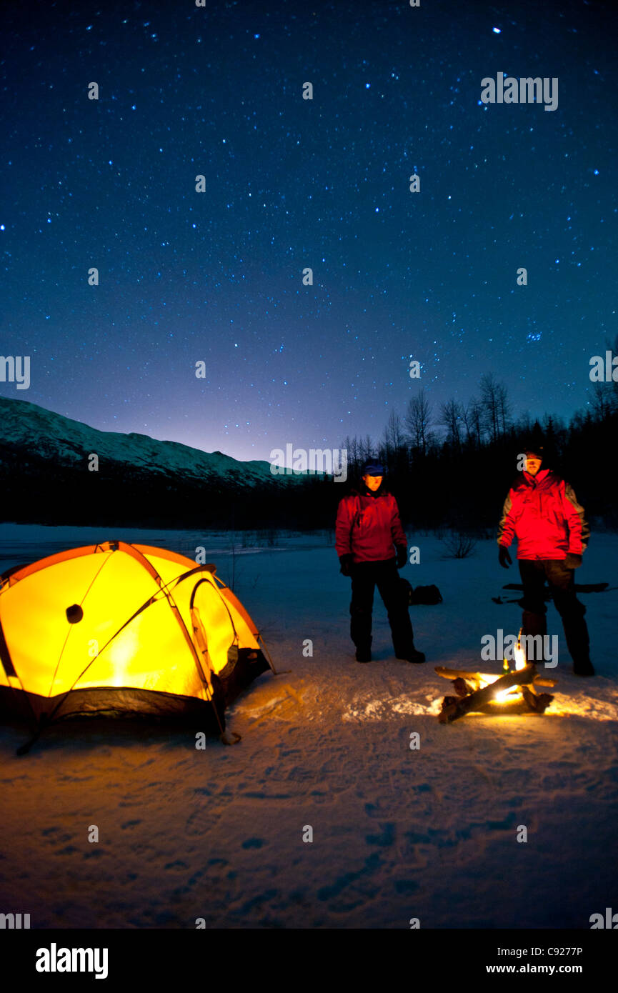 Two men warm themselves at a campfire while standing next to their lighted tent, Eklutna Lake in the Chugach State Park, Alaska Stock Photo