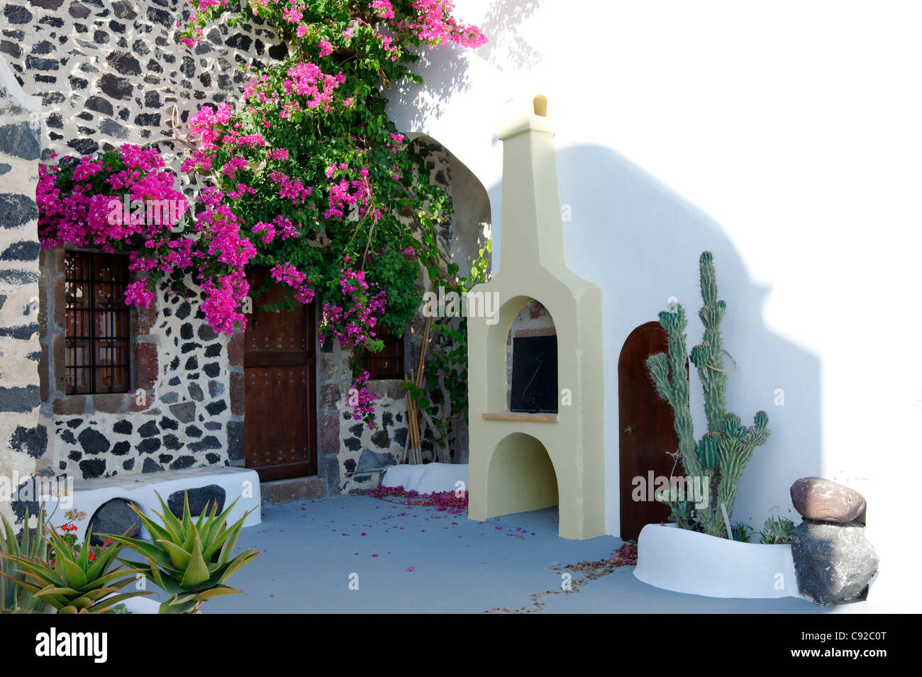 Charming view of a whitewashed courtyard wall with cacti and a Santorini rustic stone house draped in vivid pink bougainvillea. Stock Photo