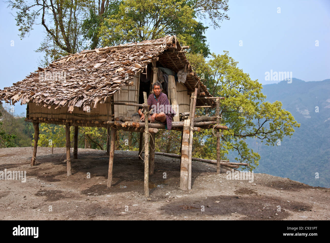 A man sits on the veranda of his small house, raised on stilts, situated precariously on the edge of a steep drop Stock Photo
