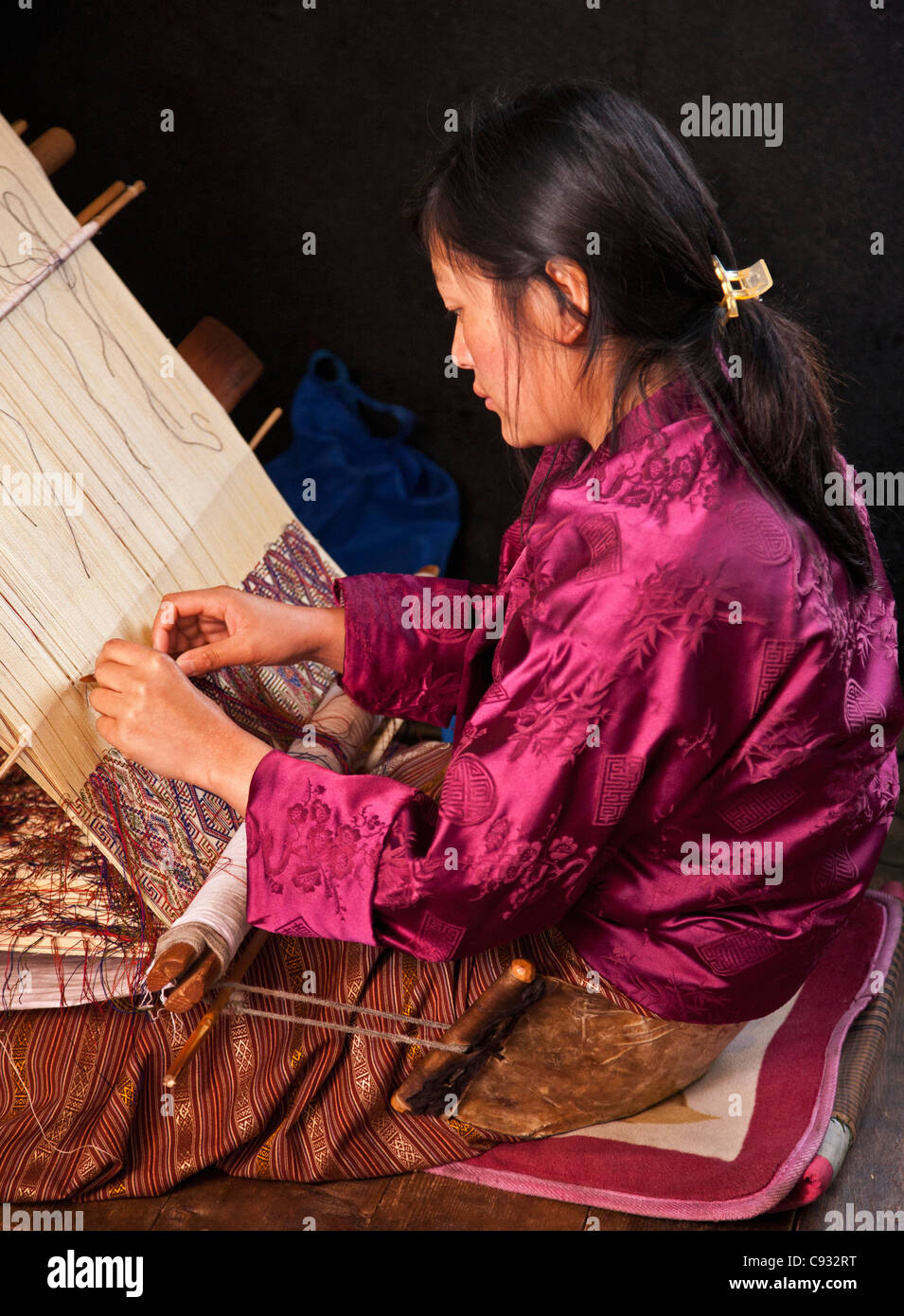 A woman weaves an intricate pattern in silk on her traditional wooden loom. Stock Photo