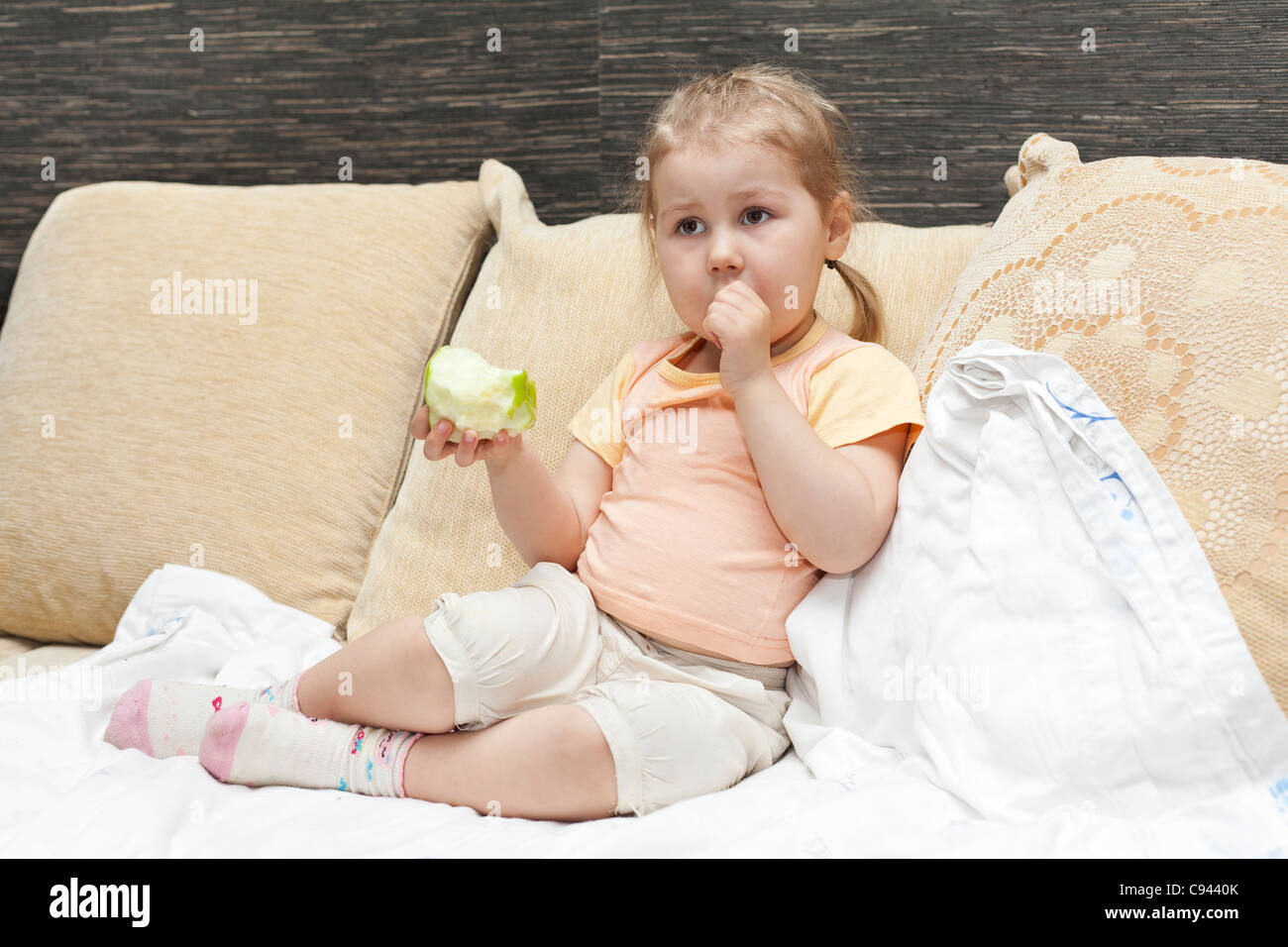 Little Russian girl sitting on sofa in domestic room and eating an apple Stock Photo