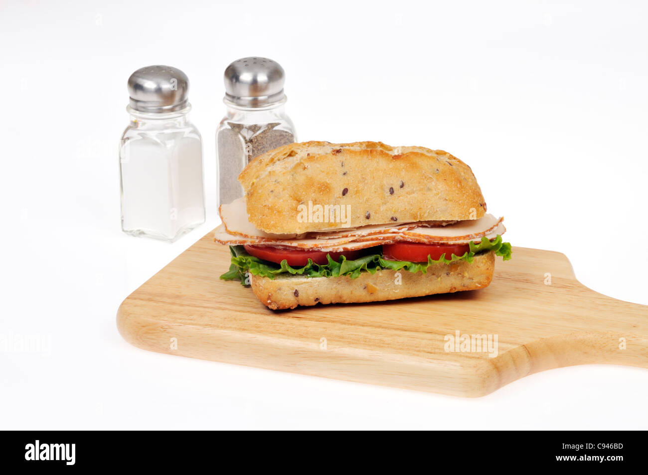 Chicken sandwich with lettuce and tomato on ciabatta bread roll with salt and pepper shakers on wood chopping board on white. Stock Photo