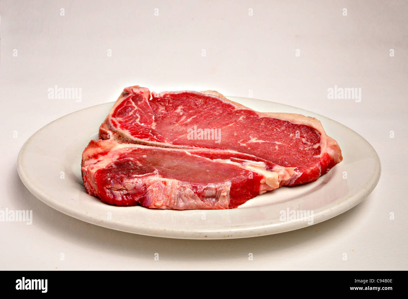 A raw uncooked t-bone steak is laying on a white plate on a plain background. Stock Photo