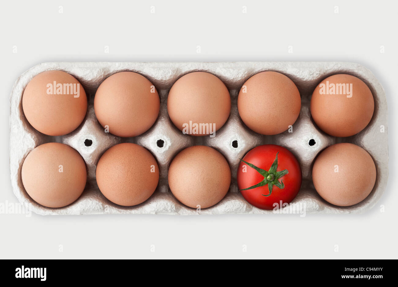 Cardboard box containing nine brown hen eggs and one single red tomato, signifying an Odd One Out concept Stock Photo
