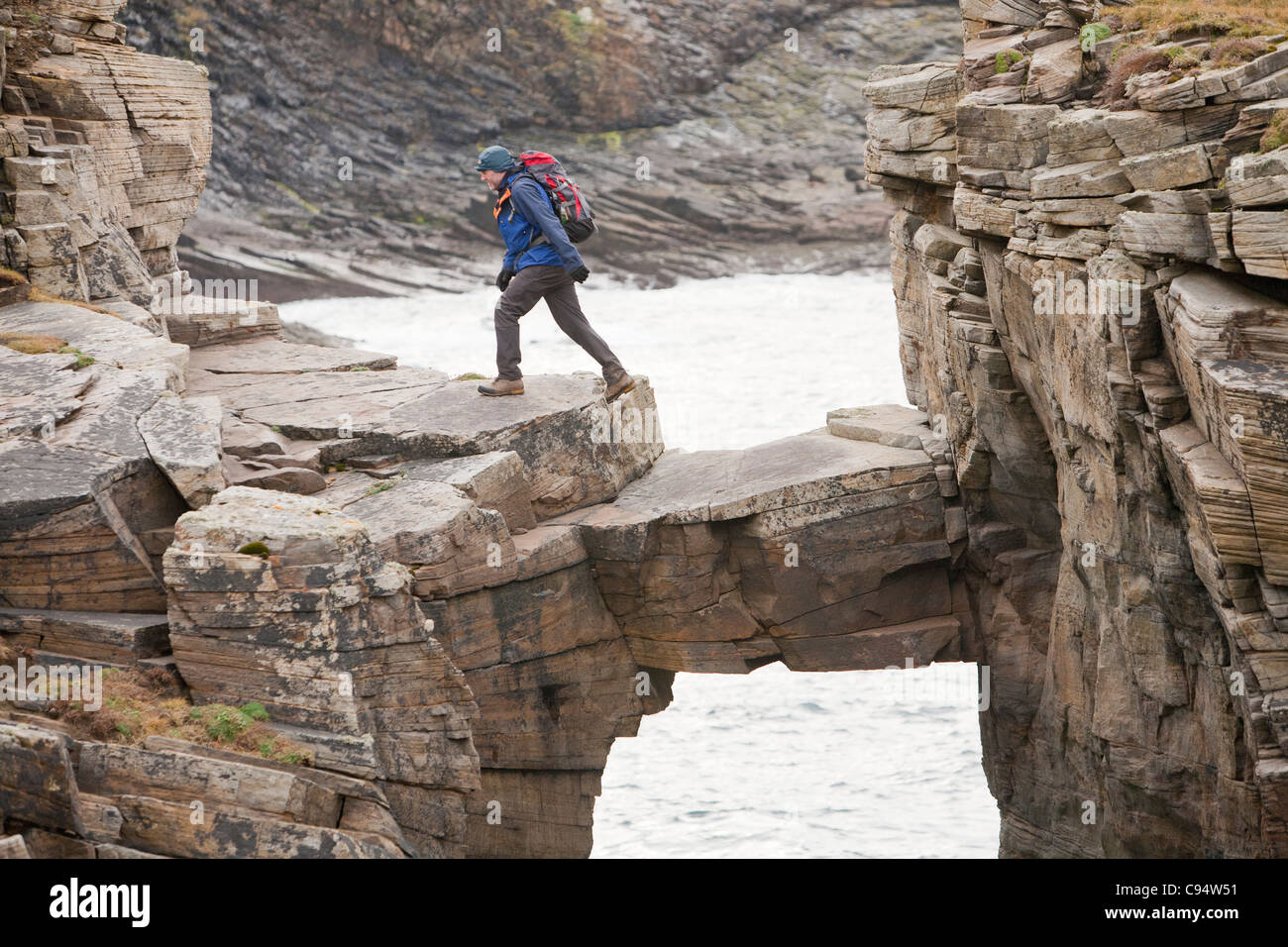 A mountaineer crosses a chock stone bridge that links the sea cliffs to a sea stack at Yesnaby on the west coast of Orkney Stock Photo