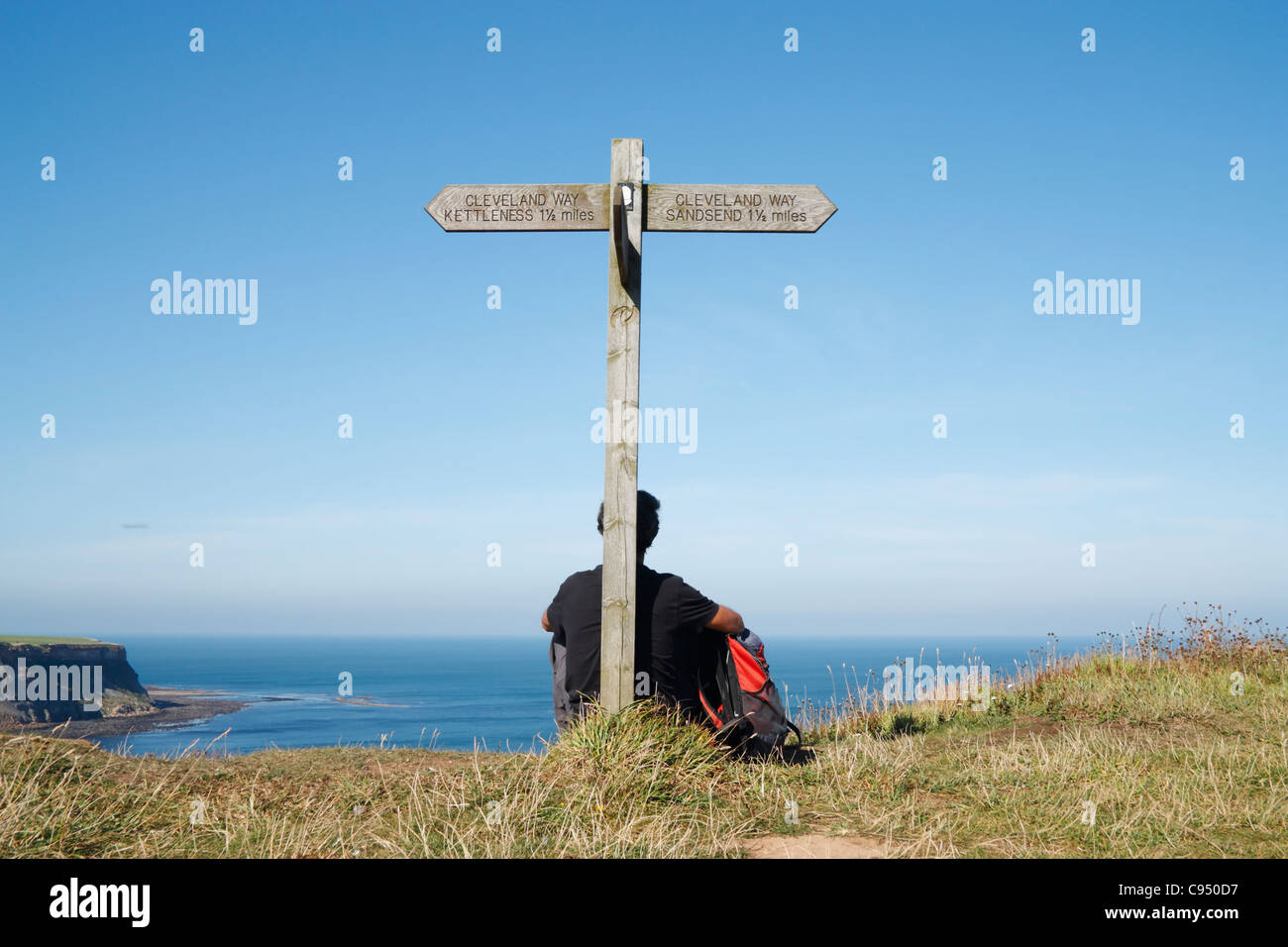 Male hiker with small rucksack sitting against signpost on the Cleveland Way coastal footpath between Kettleness and Sandsend. Stock Photo