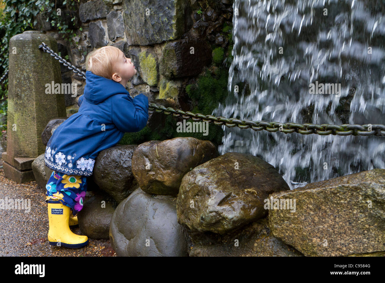 Small child in blue coat and yellow wellington boots leaning on a chain and gazing intently at an artificial waterfall. Stock Photo