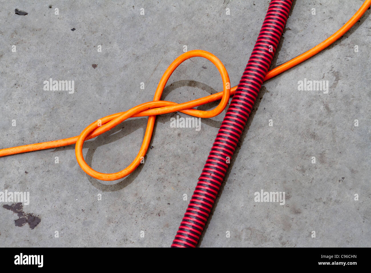 An orange electric flex with a knot in it crossed by a red and black striped plastic hose or conduit Stock Photo