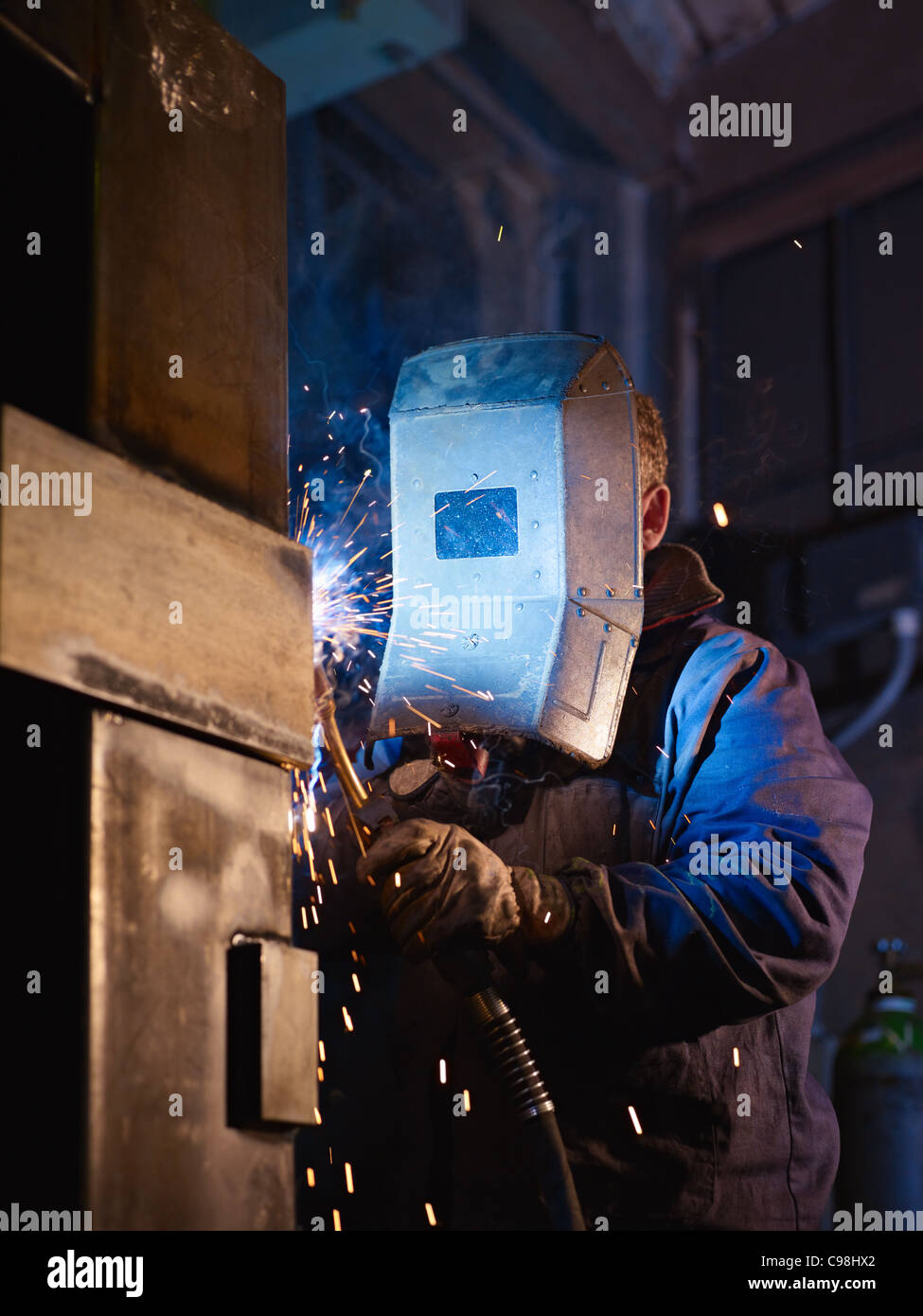 Manual worker in steel factory using welding mask, tools and machinery on metal. Vertical shape, side view, waist up Stock Photo